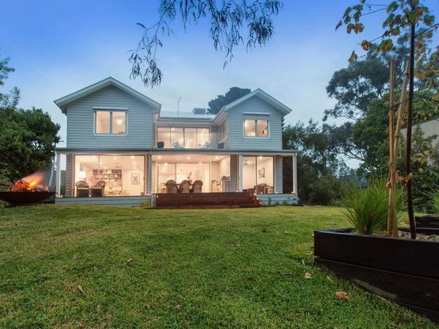 A two-storey residential house viewed from the landscaped lawn at dusk. The light-coloured weatherboard facade has two gabled upper-level wings connected by a glazed central section, with a broad covered lower-level veranda or glazed living area opening toward the garden. Warm interior lighting, timber decking, raised garden planters, mature vegetation and a small outdoor fire create an inviting setting.