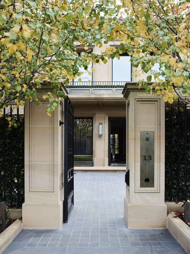 Formal gated building entrance framed by leafy trees, with pale stone-clad entry piers, black metal gates and fencing, grey tiled paving, a recessed portico, and a visible intercom panel marked 13. The entry leads toward a refined multi-storey building facade.