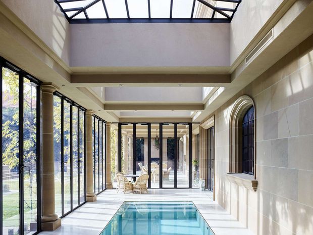A bright, glazed indoor swimming pool hall or conservatory with a long rectangular pool, full-height black-framed glazing, classical stone-clad columns, arched window detailing, and a partially glazed roof. The space combines formal classical architectural elements with a light, airy contemporary enclosure and overlooks landscaped grounds.