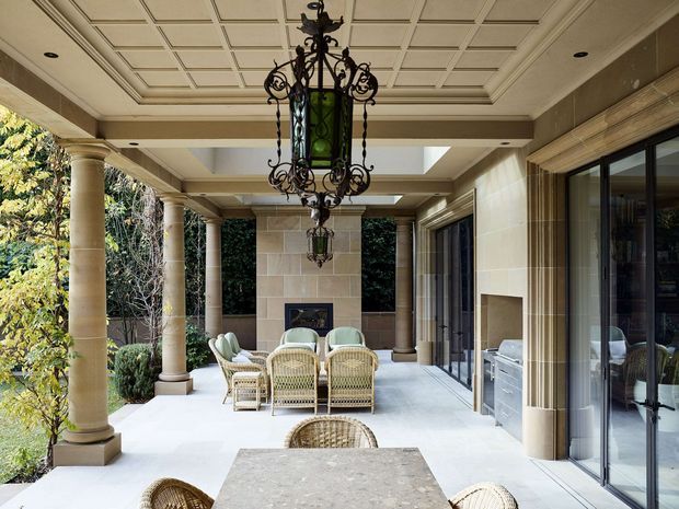 A covered outdoor terrace or loggia with classical architectural detailing, including round stone columns, a coffered ceiling, ornate wrought-iron lanterns, large glazed doors, and a built-in stone fireplace. Wicker dining and lounge furniture surround a stone-topped table, with landscaped garden visible along the open side. The space feels refined, warm, and resort-like.