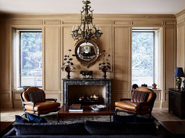 Formal traditional living room with extensive warm timber wall panelling, a carved dark stone fireplace, ornate chandelier, decorative round mirror, and two leather armchairs arranged around a central coffee table. The room has a refined, richly detailed and symmetrical character.