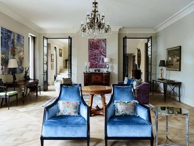 Elegant formal living room with a symmetrical arrangement of two deep-blue upholstered armchairs, a round wooden occasional table, a purple sofa, and a mirrored side table. The room features pale walls with ornate crown moulding, parquet flooring, black-framed glazed double doors, a carved timber console, framed artwork, table lamps, and a prominent crystal chandelier. The overall presentation is refined and richly decorated with a classic architectural shell and eclectic furnishings.