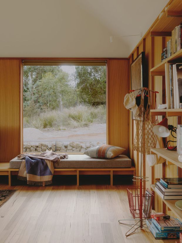 A warm, timber-lined reading nook with a built-in window seat overlooking native vegetation. The room features vertical wood wall panelling, pale timber flooring, extensive built-in shelving filled with books, and a low upholstered bench with cushions and a draped blanket. A small desk lamp, wire storage rack, and hanging textile add lived-in character. The composition has a calm mid-century-inspired feel with strong natural light and a seamless connection to the landscape.