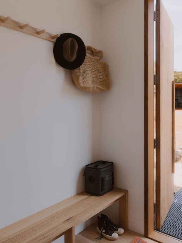 A compact, minimalist entryway viewed from inside, featuring a white plastered wall with a simple light timber peg rail holding a woven natural-fibre bag and hat. A built-in light timber bench provides seating and open shoe storage below. A tall timber-framed glazed door opens to the exterior, bringing in warm natural light.