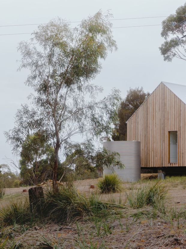 Exterior view of a compact timber-clad building set within a native, lightly landscaped rural setting. The structure has a steep gabled roof, vertical natural-toned timber cladding, a small vertical window, and a corrugated metal cylindrical water tank beside it. Sparse grasses, eucalyptus-like trees, sandy ground, and overhead utility wires contribute to an informal Australian bush or rural character.