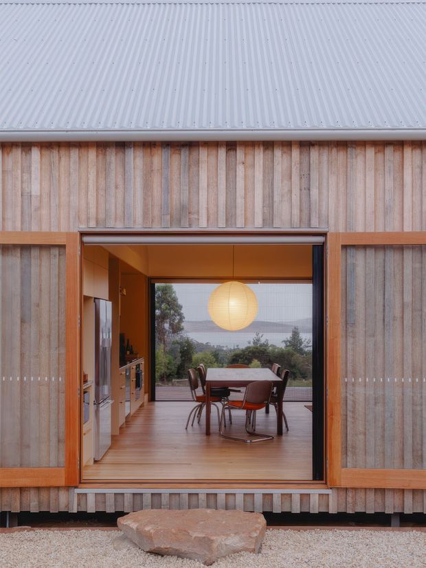 A straight-on exterior view of a compact timber-clad building with a pale corrugated metal roof and large open sliding doorway. Through the opening, a warm timber-lined dining area and adjacent kitchen are visible, furnished with a rectangular dining table, chairs, and a large spherical pendant light. The glazing beyond frames a distant natural landscape. A rough stone stepping slab sits in a pale gravel foreground. The composition is simple and contemporary, with strong indoor-outdoor connection and natural timber tones.