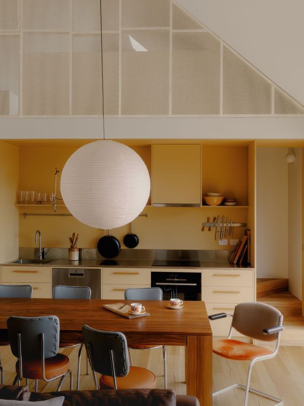 Warm, compact open-plan kitchen and dining area with mustard-yellow built-in cabinetry and wall finish, timber dining table, mid-century style seating, stainless-steel appliances, and a large white spherical pendant light. A screened mezzanine or stair landing with a sloped underside is visible above the kitchen, while timber flooring continues into the adjacent space.