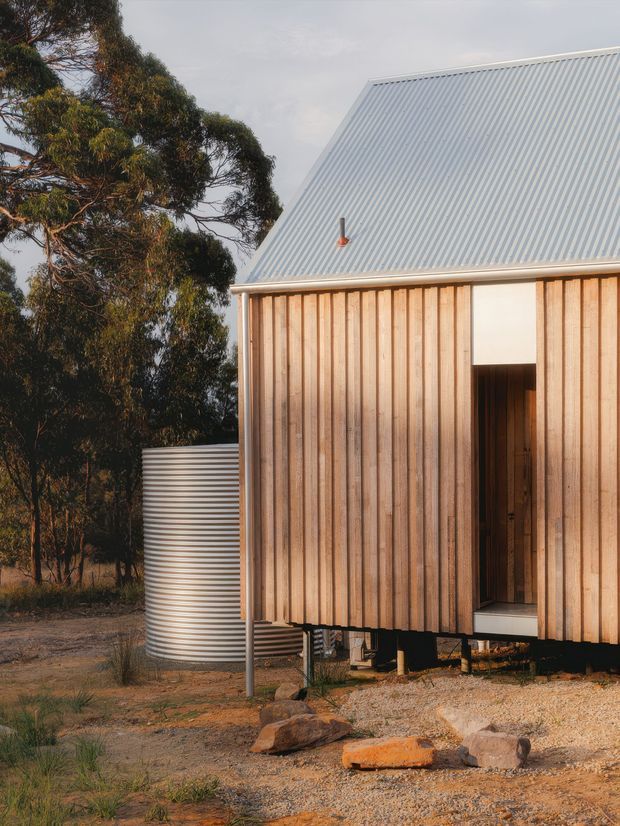 Exterior view of a compact timber-clad building set in a natural bushland setting. The structure has vertical light timber boards, a steep simple gable roof clad in corrugated metal, a recessed dark doorway, and a cylindrical corrugated water tank beside it. The building appears elevated slightly above the rocky, sparsely vegetated ground.