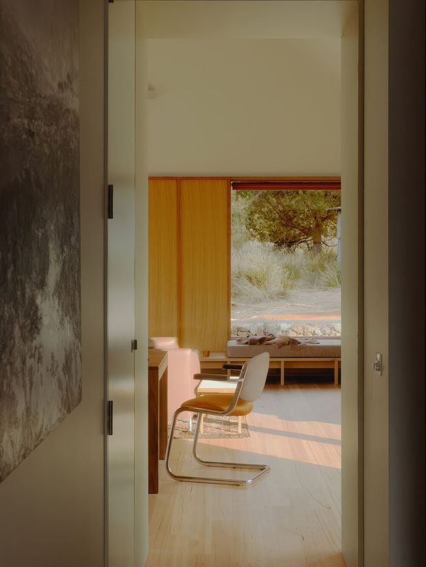 View through an open doorway into a warm, sunlit interior workspace or dining area. The room has pale timber flooring, a timber-framed window with a low built-in bench, warm timber wall panelling, and a modern cantilever-style chair positioned at a desk or table. A large textured wall artwork or feature surface is visible in the foreground, while dry grasses and trees can be seen outside.