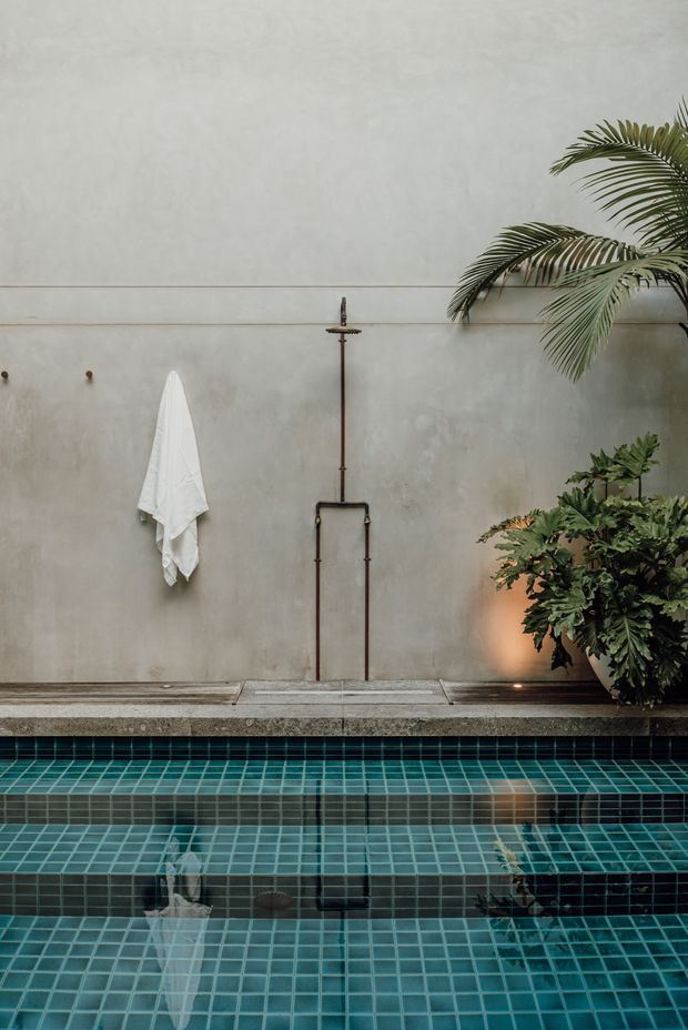 A tranquil courtyard pool scene with a turquoise small-square tiled pool in the foreground, a mottled grey plaster wall behind it, exposed dark metal pool plumbing and shower fixture, a white towel hanging from a wall hook, and lush tropical foliage including a palm. Warm recessed lighting highlights the plants and creates reflections across the still water. The composition is minimalist and spa-like.