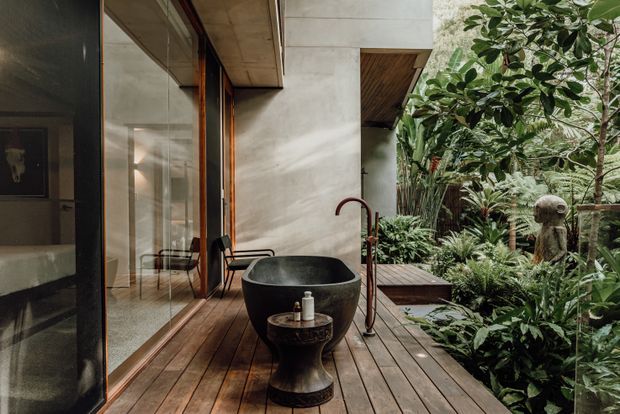 A moody, tropical outdoor bathroom or bathing area on a timber deck, with a freestanding dark vessel bathtub, matching pedestal basin, and exposed copper-toned plumbing. Floor-to-ceiling glazing opens toward a lush planted courtyard, while concrete walls, timber soffits, and timber decking create a sheltered indoor-outdoor setting. A sculptural stone figure is visible among the foliage.