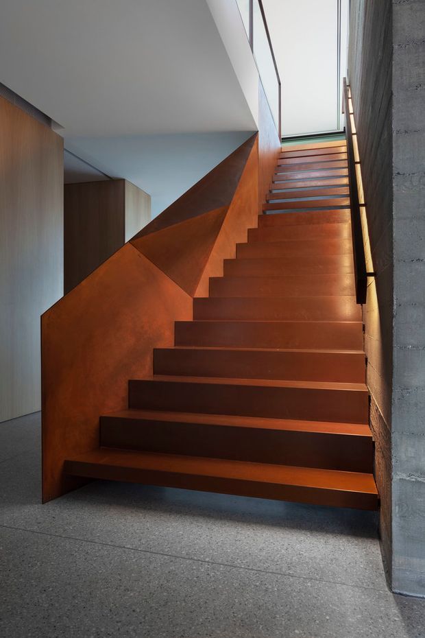 A contemporary interior stairway with a sculptural, angular rust-brown metal staircase rising beside a rough exposed concrete wall. The stairs are illuminated by a tall glazed skylight or clerestory, with a slim black handrail and warm timber-toned wall surfaces. The floor is grey polished concrete or terrazzo.