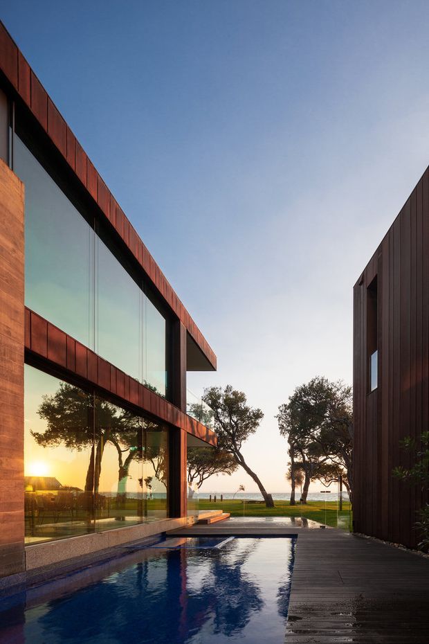 A modern coastal residence is framed around a narrow reflective swimming pool and timber deck. The building features dark vertical cladding, expansive floor-to-ceiling glazing, strong horizontal cantilevered forms, and warm rust-toned metal cladding. Coastal vegetation and the ocean are visible beyond the property, with low evening sunlight reflected in the glass and water.