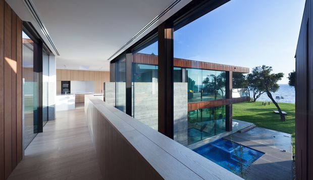 A bright contemporary interior corridor overlooking a landscaped waterfront property and swimming pool. The corridor features extensive dark-framed floor-to-ceiling glazing, warm timber wall and cabinetry finishes, pale timber flooring, and a long built-in ledge. A minimalist kitchen is visible at the far end, while a modern glazed building wing, lawn, trees, and pool are seen outside.