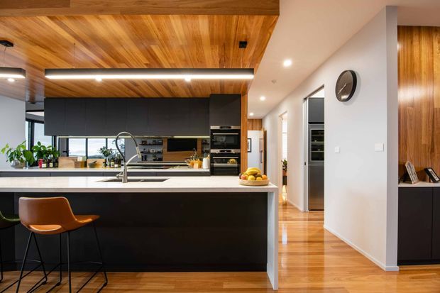 A contemporary open-plan kitchen dominated by a large black island with a white benchtop and integrated sink. Dark cabinetry, built-in appliances, timber flooring, and a striking timber-lined ceiling create a warm, refined contrast. The kitchen opens toward a bright living or dining area, with a hallway and additional built-in joinery visible to the right.