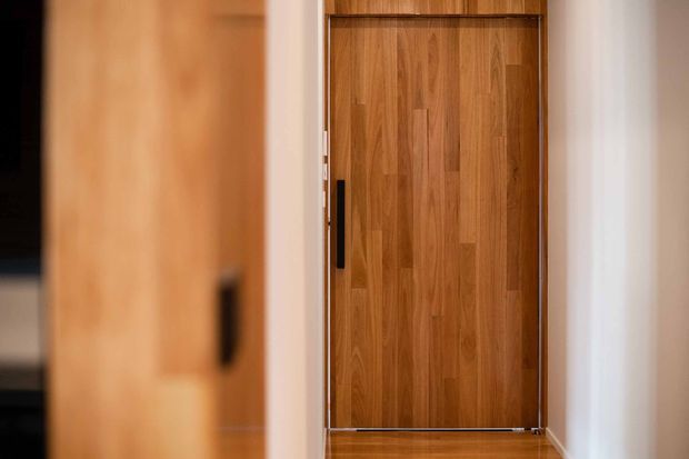 A close view down a narrow interior passage toward a tall, richly grained timber door or concealed joinery panel. The door has a slim dark vertical handle and dark edging, framed by light neutral walls and warm timber surfaces. The foreground includes a softly blurred timber panel, creating a shallow-depth-of-field architectural detail photograph.