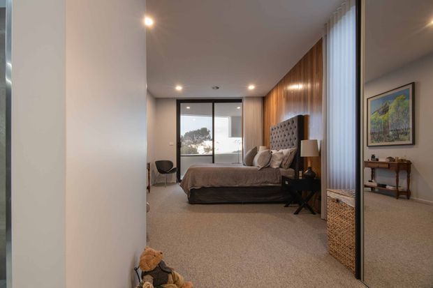 A modern bedroom viewed from the entry, featuring a low-profile upholstered bed with a tufted headboard, warm vertical timber wall panelling, neutral carpet, recessed ceiling lights, full-height windows with vertical blinds, and a large mirrored wardrobe or wall panel. A small side table, lamp, chair, woven storage basket, and framed artwork complete the room.