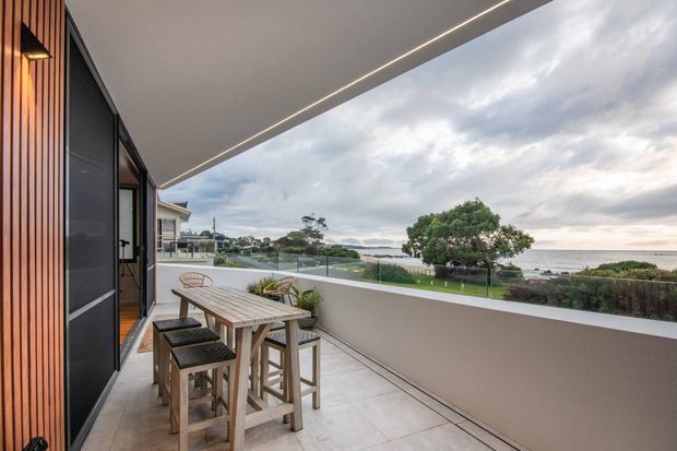 A contemporary covered terrace or balcony overlooking a coastal landscape. The space features a long timber outdoor dining table with bar-height stools, pale tiled flooring, a solid rendered balustrade topped with clear glass, dark framed glazing, and a vertical timber-clad wall with integrated lighting. The broad roof overhang and linear recessed lighting create a sheltered, refined outdoor entertaining area with expansive ocean views.
