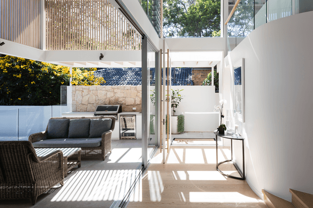 Bright contemporary indoor-outdoor living space with a glazed connection to a covered patio and outdoor entertaining area. The interior has pale timber flooring, white walls, extensive glazing, a slim black-framed glass door, and a high ceiling with timber screening. A rattan lounge setting occupies the adjacent outdoor area, while the patio includes a built-in barbecue, stone wall, plants, and a simple console table. Strong linear sunlight and shadow patterns create an airy, refined atmosphere.