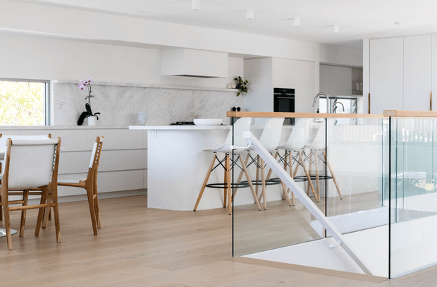 Bright open-plan contemporary kitchen and dining area viewed from a stair landing. The space features white full-height cabinetry, a white island with a pale stone benchtop, a marble-look splashback, pale timber flooring, white bar stools, and a glass balustrade enclosing the stairwell. Natural light enters through a large window, creating an airy, clean interior.