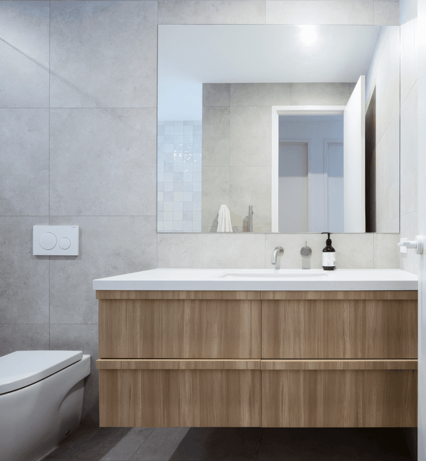 A clean, contemporary bathroom vanity viewed straight on. The room features large light-grey wall tiles, a large frameless mirror, a white integrated basin and benchtop, warm timber-grain vanity cabinetry, dark floor tiles, and a partially visible wall-hung toilet. A simple chrome tap and dark soap dispenser sit on the vanity.