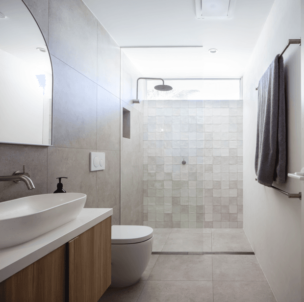Bright contemporary bathroom with a wall-mounted rainfall shower, frameless glass shower enclosure, pale square tiled feature wall, large-format grey floor and wall tiles, floating timber vanity with white vessel basin, wall-hung toilet, and a heated towel rail with dark towel.