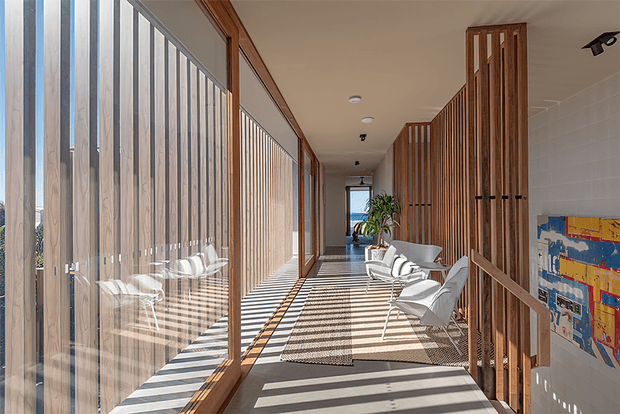 A bright, elongated interior corridor or gallery-like hallway with full-height glazing and vertical timber screening along the left side, casting strong striped shadows across the floor. Timber slatted partitions and doors line the right side, with a small seating area of white lounge chairs, a neutral rug, artwork, and indoor planting. The space has a warm, refined contemporary character with strong indoor-outdoor visual connection.