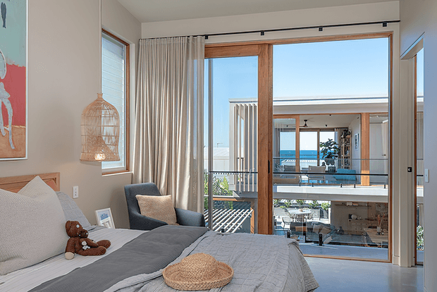A bright, contemporary bedroom with a neatly made bed, neutral grey and cream bedding, a blue upholstered armchair, and a woven pendant light. Full-height timber-framed glazing and a large opening provide views into a modern multi-level living space with a balcony and abundant natural light. The room has a calm, refined coastal-contemporary feel.