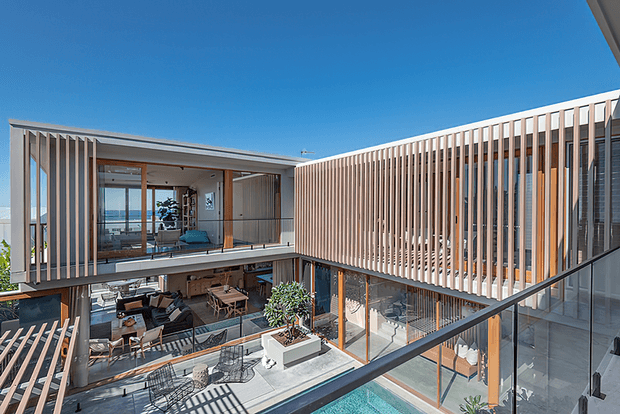 Contemporary multi-level courtyard residence viewed from above, featuring extensive glazing, warm timber vertical screening, open-air terraces, outdoor dining and living areas, and a visible swimming pool. The design creates a sheltered, light-filled courtyard with strong indoor-outdoor connections.