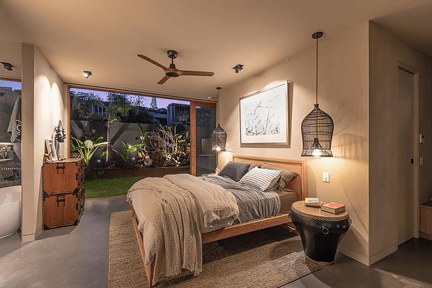 Warm, contemporary bedroom with a low timber bed, neutral linen bedding, a textured area rug, pendant lighting, ceiling fan, framed artwork, and a timber-framed opening to a planted courtyard. A bathroom is partially visible through the opening on the left.