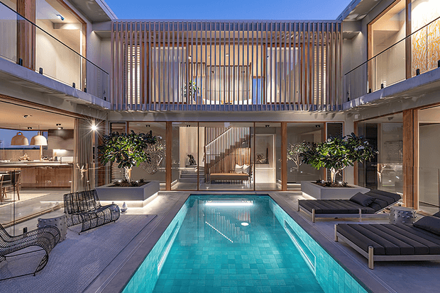 A sophisticated indoor courtyard centered around a long rectangular swimming pool, surrounded by timber decking, lounge furniture, landscaped planters, and glass-lined living spaces. The double-height atrium features warm timber slatted screening, exposed concrete elements, extensive glazing, and a visible upper-level walkway, creating a refined indoor-outdoor resort atmosphere.