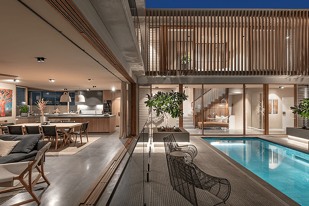 A sophisticated open-plan interior centred around an indoor swimming pool and double-height void. The space combines a lounge, dining area and kitchen with warm timber cabinetry, exposed concrete, dark metal framing, extensive glazing and vertical timber screening around an upper-level mezzanine. The poolside terrace includes wire-frame lounge chairs, while large indoor plants soften the contemporary, refined setting.