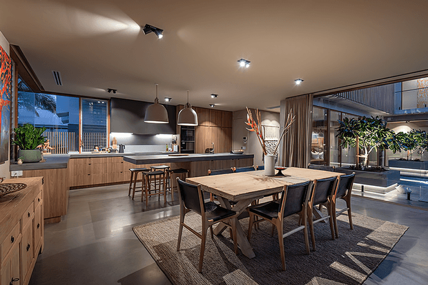 An open-plan contemporary kitchen and dining area with extensive timber cabinetry, a dark island bench with bar stools, and a large timber dining table surrounded by upholstered chairs. Floor-to-ceiling glazing and sliding doors open toward a planted courtyard or outdoor area, while track and pendant lighting create a warm, refined atmosphere.