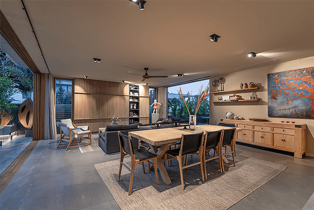 A spacious contemporary open-plan living and dining area with a large timber dining table, upholstered dining chairs, low lounge seating, built-in timber wall cabinetry, floating shelves, artwork and expansive glazed openings. The room has polished concrete flooring, warm timber panelling, recessed and track lighting, and views to lush outdoor planting.