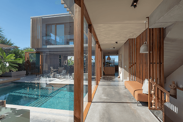 A bright contemporary residential corridor beside a swimming pool, with full-height glazing framed in warm timber, polished concrete flooring, vertical timber screening, and a stairway along the right wall. The view extends toward a kitchen or living area, while a modern two-storey building and landscaped pool terrace are visible outside.