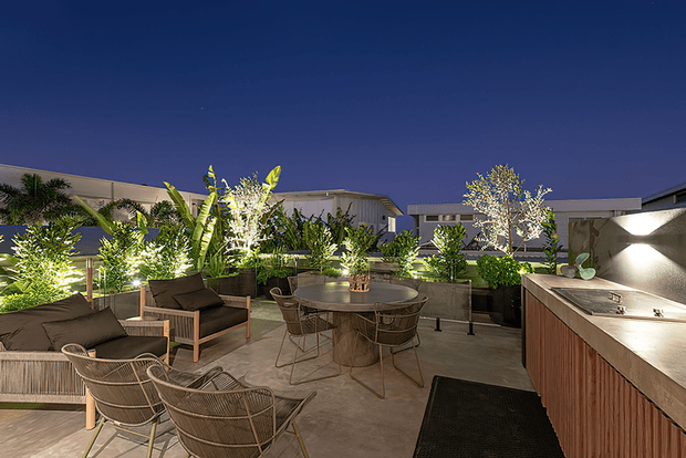 A contemporary rooftop or elevated outdoor dining and lounge terrace photographed at dusk. The space features a concrete-paved floor, a large dining table with wire-frame chairs, woven lounge chairs and sofas, raised planter beds filled with lush tropical and ornamental planting, and integrated exterior lighting. A timber-clad outdoor kitchen or bar counter occupies the right side, with low modern buildings visible around the perimeter.