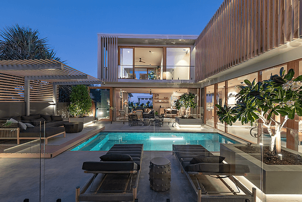 A sophisticated multi-level contemporary residence arranged around a turquoise swimming pool and outdoor entertaining area. The image shows large glazed openings, warm timber vertical screening, concrete-toned paving, integrated planting, lounge furniture, sun loungers, and a covered outdoor dining space. The design creates a private courtyard-like resort atmosphere with strong indoor-outdoor connection.