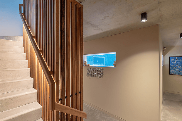 Interior view of a contemporary stairway with pale concrete stairs, a timber vertical-slat balustrade and handrail, and a warm neutral hallway. A small blue-tinted window, recessed ceiling lights, polished concrete flooring, and artwork are visible.