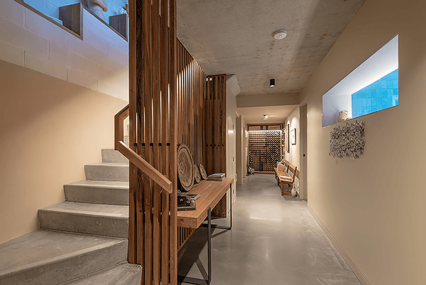 A refined contemporary interior hallway with a polished concrete floor and ceiling, warm cream walls, and a sculptural open timber staircase. Vertical timber slats form a partition and balustrade around a slim console table displaying decorative objects. The corridor extends toward a patterned screen, with recessed wall niches, bench seating, artwork, and a glowing blue-toned feature opening. The space combines warm timber detailing with minimalist concrete finishes and soft indirect lighting.