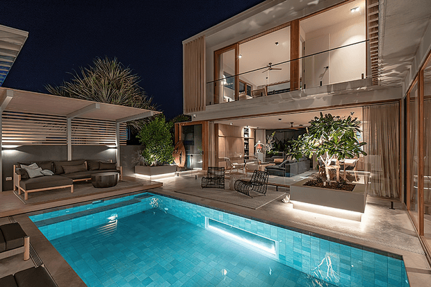 Night-time view of a contemporary two-storey residence centred around a brightly illuminated swimming pool. The scene includes extensive floor-to-ceiling glazing, warm timber screening and cladding, a covered outdoor lounge with slatted roof, landscaped planters, and an open-plan interior visible through the glass.
