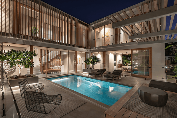 A sophisticated two-storey courtyard residence at night, centred around a rectangular illuminated swimming pool. The open-plan outdoor living area features timber decking, dark woven lounge chairs, a low round fire-pit or table, large planters, extensive glass walls, and a dramatic timber batten privacy screen. Exposed structural beams and pergola-like roof framing define the upper level and create a refined indoor–outdoor connection.