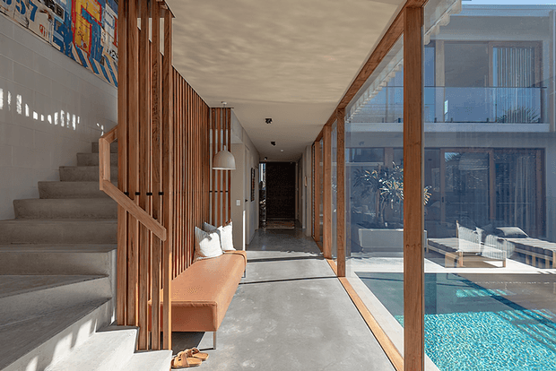 A bright contemporary residential hallway with a central concrete floor, a staircase on the left screened by vertical timber battens, and full-height timber-framed glazing on the right overlooking a turquoise swimming pool and an adjacent modern building. A built-in-style upholstered bench sits beneath a pendant light, with artwork and concealed dark joinery at the far end.
