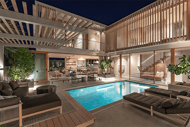 Luxurious multi-level indoor courtyard with a central swimming pool, timber decking, extensive vertical timber screening, open-plan living and dining areas, indoor plants, and a mezzanine walkway. The double-height space is warm and sophisticated, with strong indoor-outdoor resort-like character and integrated lounge areas around the pool.