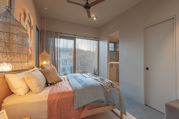 A warm, contemporary bedroom with a neatly made bed layered in white, blush, and grey textiles. The room features full-height vertical blinds, a large woven pendant light, a decorative wire birdcage-style wall light, timber accents, a ceiling fan, and a modern flush-panel door. Soft evening lighting creates a cozy atmosphere.