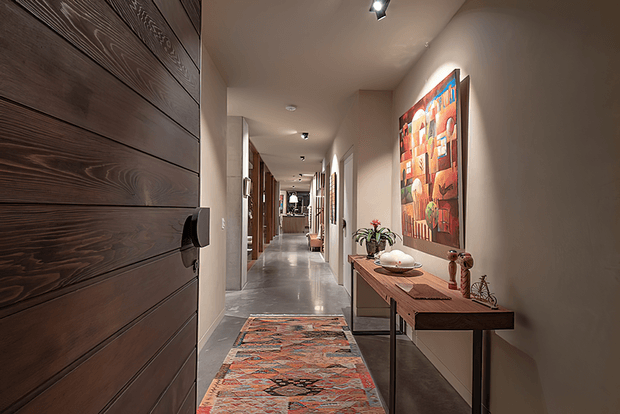 A long, warmly lit residential hallway with polished grey flooring, cream walls, and a dark horizontal timber-panelled door in the foreground. A patterned runner leads toward multiple doorways and recessed ceiling lights. On the right, a narrow timber console displays decorative objects and a plant beneath a colourful abstract artwork.