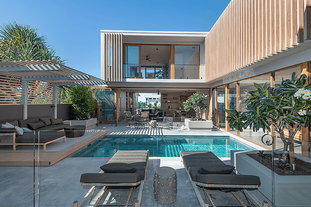 A bright contemporary courtyard centred on a rectangular swimming pool, with a two-storey residence surrounding the space. The building features extensive floor-to-ceiling glazing, warm vertical timber cladding, white structural framing, and a covered outdoor lounge with slatted screening. Poolside loungers, built-in seating, planters, and mature greenery create a refined indoor–outdoor entertaining area.