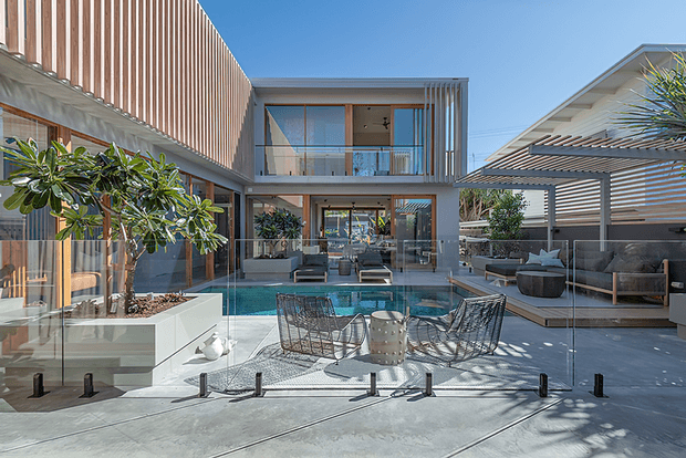 A bright contemporary multi-level residential courtyard viewed from an outdoor entertaining area, centred on a rectangular swimming pool. The home features extensive glazing, timber vertical screening, concrete paving, glass pool balustrades, and a covered outdoor lounge with a slatted roof. Outdoor seating and planters surround the pool in a refined modern setting.