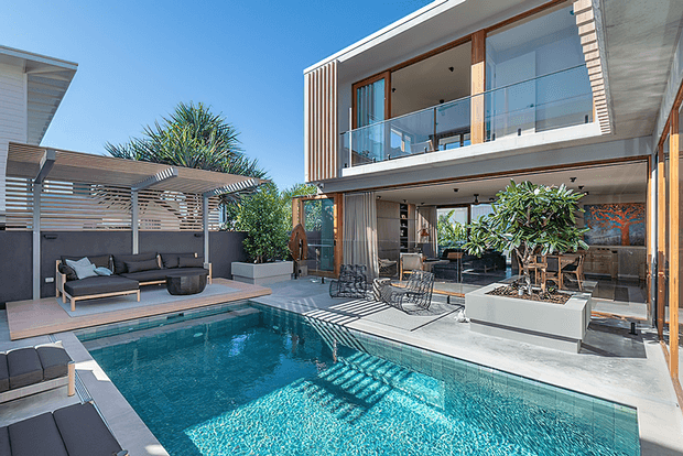 Contemporary two-storey residence with extensive glass walls and balconies opening onto a landscaped pool terrace. The outdoor area features a rectangular swimming pool, timber-toned decking, dark outdoor lounge seating beneath a slatted pergola, raised planters, and a covered entertaining zone.