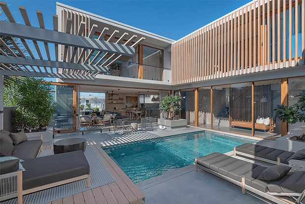 A luxurious contemporary courtyard-style residence with an indoor-outdoor living area opening onto a rectangular swimming pool. The space features extensive glazing, warm timber vertical screening, a dramatic steel pergola, grey outdoor lounge furniture, timber decking, and a bright blue sky visible above.