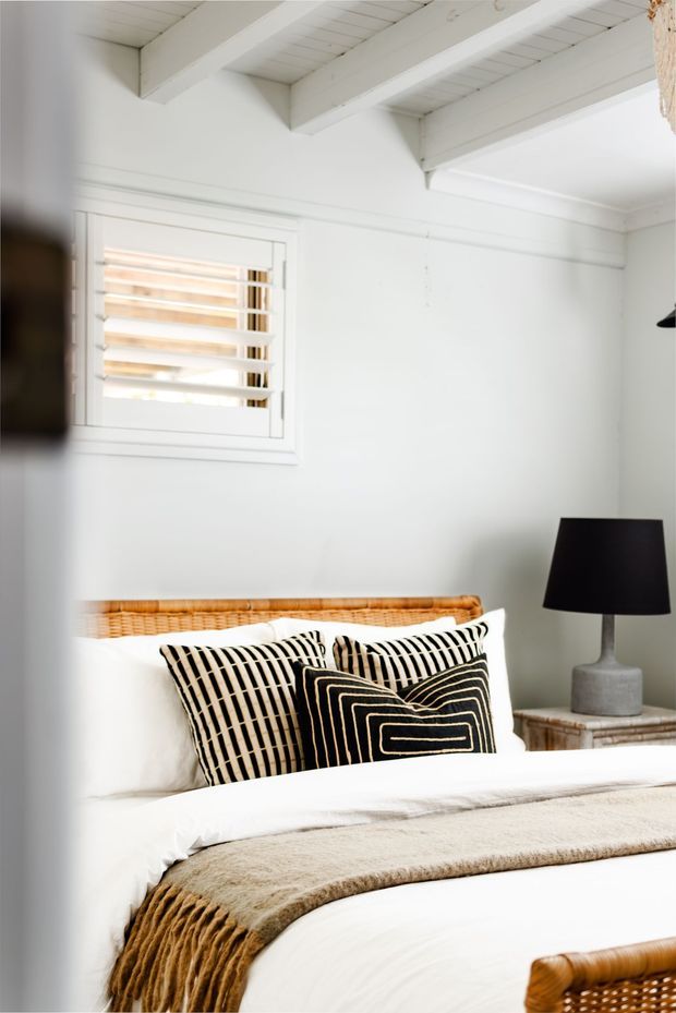 Bright, compact bedroom with white walls and exposed white-painted ceiling beams. A woven rattan headboard and matching rattan furniture are paired with crisp white bedding, patterned black-and-cream cushions, a textured beige throw, and a black table lamp on a small bedside table. A white-framed window with horizontal louvres provides natural light.